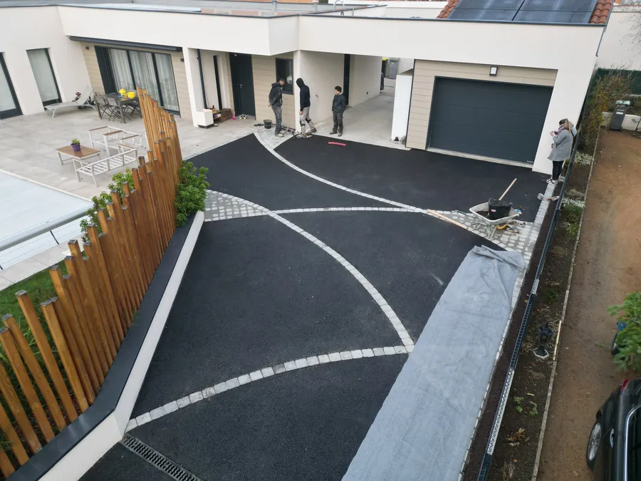 Allée en enrobé noir avec bandes de pavés arrondis devant une maison